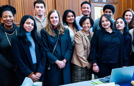 Photo of a large group of smiling people standing around a desk with a laptop computer