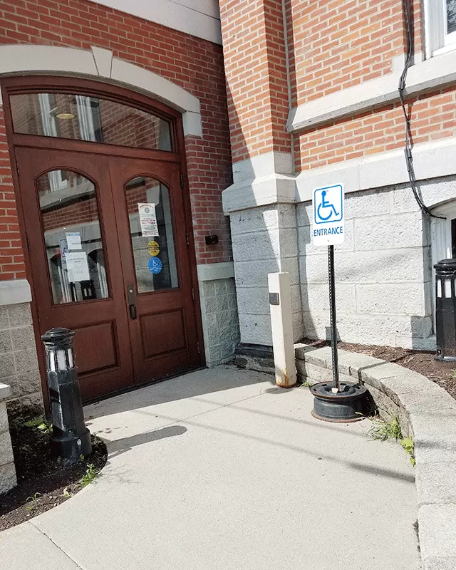 Description of photo: There is a curved walkway that leads to double doors. Within the walkway is ADA signage and a push button to open the right door.
