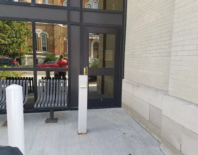 Description of photo: Courthouse with a single door to the far right. To the left is a push button that will open the door. There is outdoor seating to the left of the door and a push button.