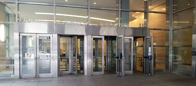 Description of photo: The multiple door entrance to Family Court from street level. Double doors marked with ADA signage to the far left, followed by a revolving door and then two additional double doors. Each of the double doors on the right have one door open.