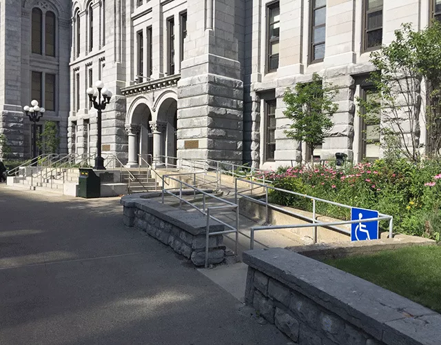 Description of photo: A set of stairs with railings leading to the entrance. There is another set of stairs to the right that also leads to the entrance. To the far right is a long ramp that directs you to the entrance.