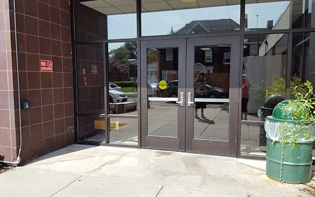 A set of double doors. To the far left against the wall is a push button to automatically open the door and a no smoking sign.
