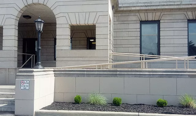  An entrance with stairs leading directly to the entry way. To the right, there is a ramp that also leads to the entry way. In front of the ramp are small shrubs.