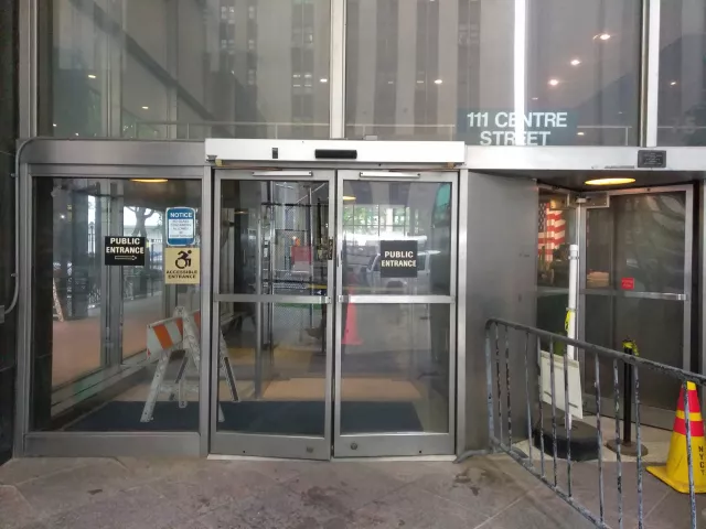 Description of photo: A double door entrance labeled Public Entrance is located on the street level. To the left of the double doors is ADA signage. There is a barricade to the right of the double door entrance.