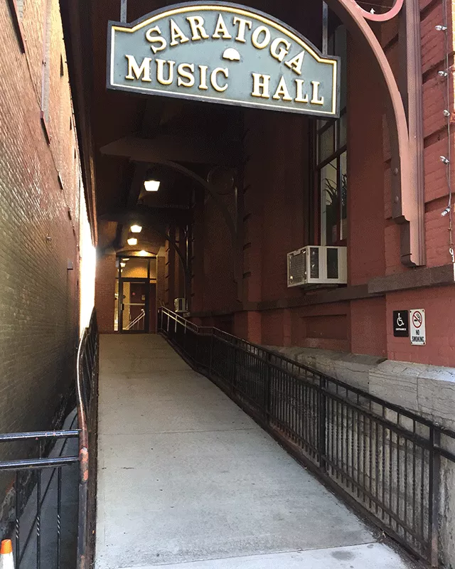 Description of photo: A ramp leading to a single door. There is ADA signage and a no smoking sign on the right hand side of the building. Above the ramp is a sign that says 'Saratoga Music Hall.'