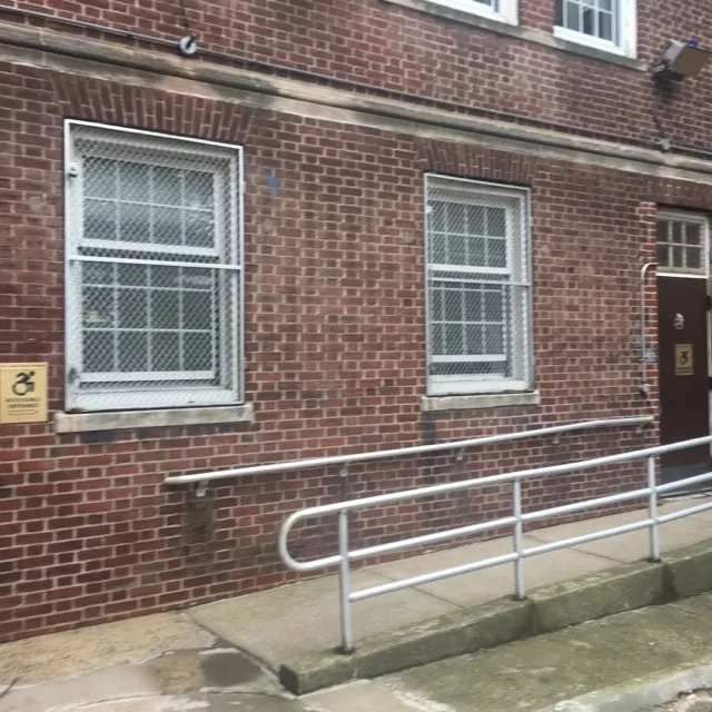 Description of photo: A ramp leading to a single door with ADA signage. Toward the entry area of the ramp, there is ADA signage posted on the side of the building.