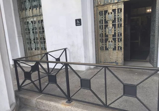 A ramped walkway leading to a set of double doors. The door to the right is open. There is signage posted on the building to the left of the doors.
