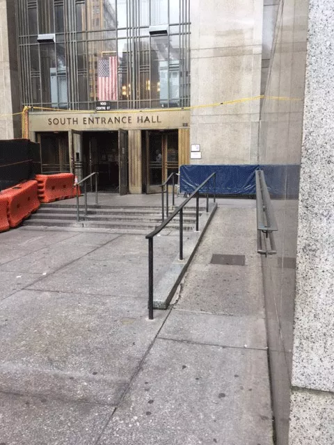 A walkway leading to steps. The steps lead to two revolving doors separated by double doors entry. The double door entry is open. To the right of the walkway is a ramp that leads to the entrance area.