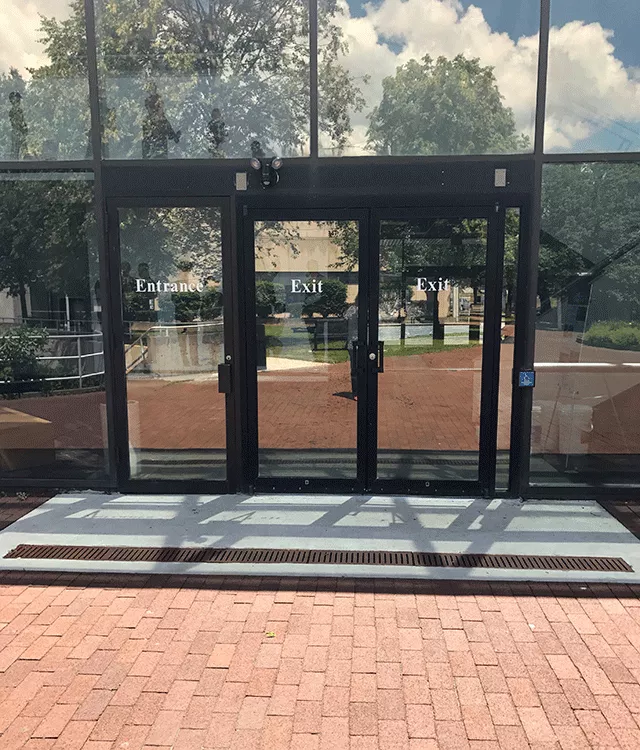 View of three doors to the court facility. The door to the left has the word "Entrance" and the two doors to the right have the word "Exit." There is a push button on the pole toward the right of the farthest right door.