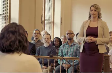 A woman stands in a courtroom with others seated behind a wooden railing
