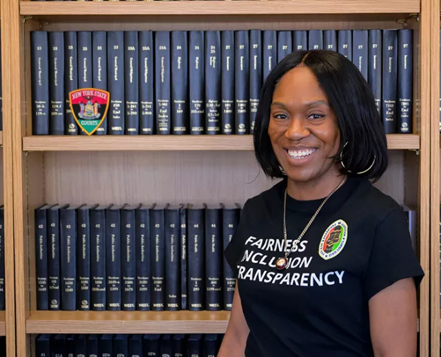 Photo: Senior Court Clerk Rachel Walcott, the creative force behind the BHM Commemorative Patch’s design, stands before a bookshelf displaying the newly unveiled patch, a symbol of unity, cultural pride, and equality. 