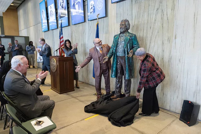 Rochester City Court Supervising Judge Barrett at the podium as Monroe County Commissioner of Jurors Perreaud, and Monroe Deputy Commissioner of Jurors Rainford-David unveil the statue bearing Mr. Douglass’s likeness 