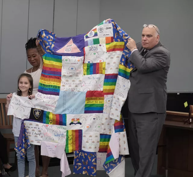 Acting Supreme Court Justice Fields (left), Law Clerk Harry Tilis (right), and 11-year-old Lily Coysh, who spoke of what “allyship” means to her, display the Suffolk County Courts’ colorful, evolving Quilt of Equality during the courthouse PRIDE event 