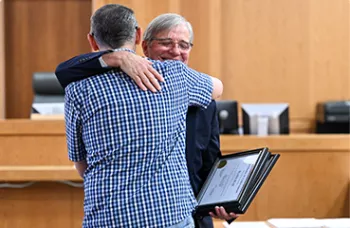 An older man smiles warmly while being hugged by another person in a courtroom