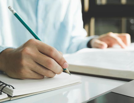 Photo of someone writing in a notebook with a pencil while referring to a book.