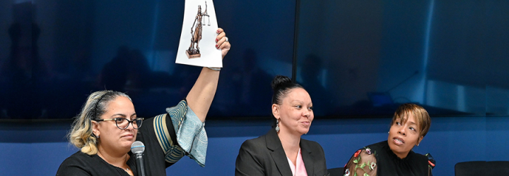 Photo of three woman sitting at a desk. One of them holds up a piece of paper with Lady Justice on it.